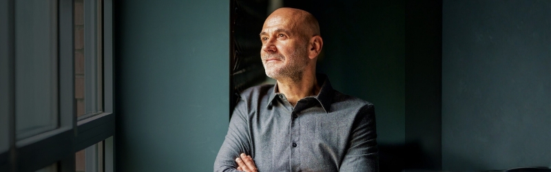 Middle aged business man in navy shirt looks into distance leaning against a turquoise wall.