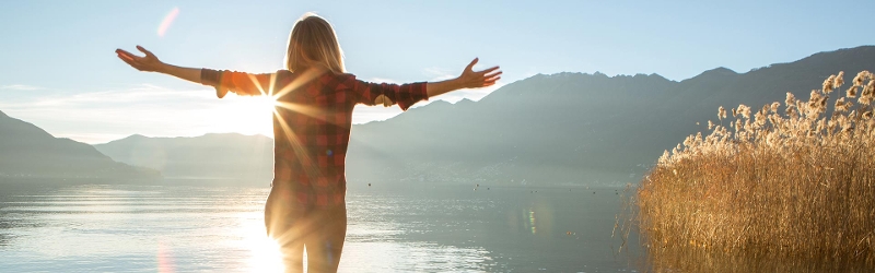 Woman standing on a rock with her arms out embracing the sun while glaring  at the lake in front