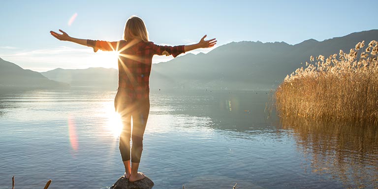 Woman standing on a rock with her arms out embracing the sun while glaring  at the lake in front